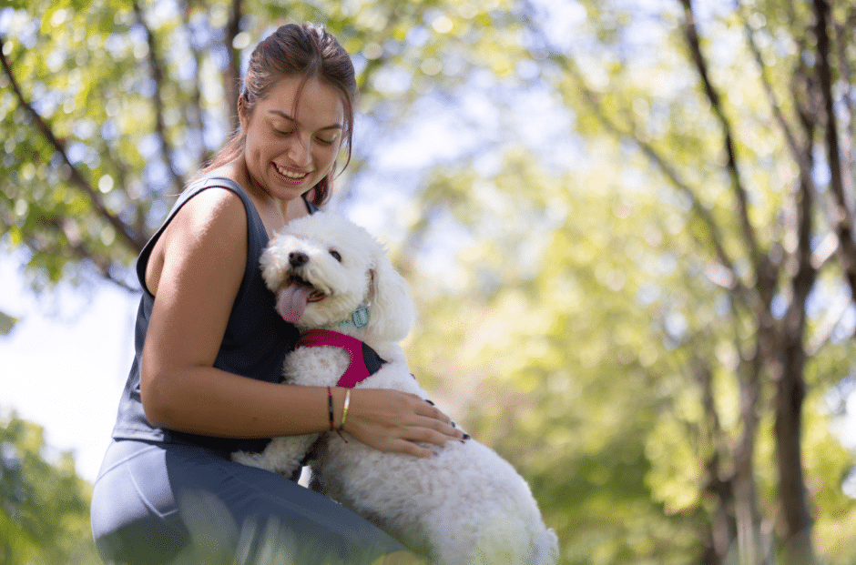 dog grooming near burnham - A woman smiling and holding a fluffy white dog wearing a pink harness in an outdoor park setting with trees in the background.