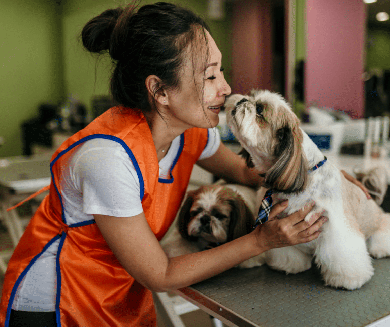 dog grooming near goldhanger - A dog groomer in an orange apron smiling and interacting affectionately with a small dog on a grooming table, with another dog resting beside them.